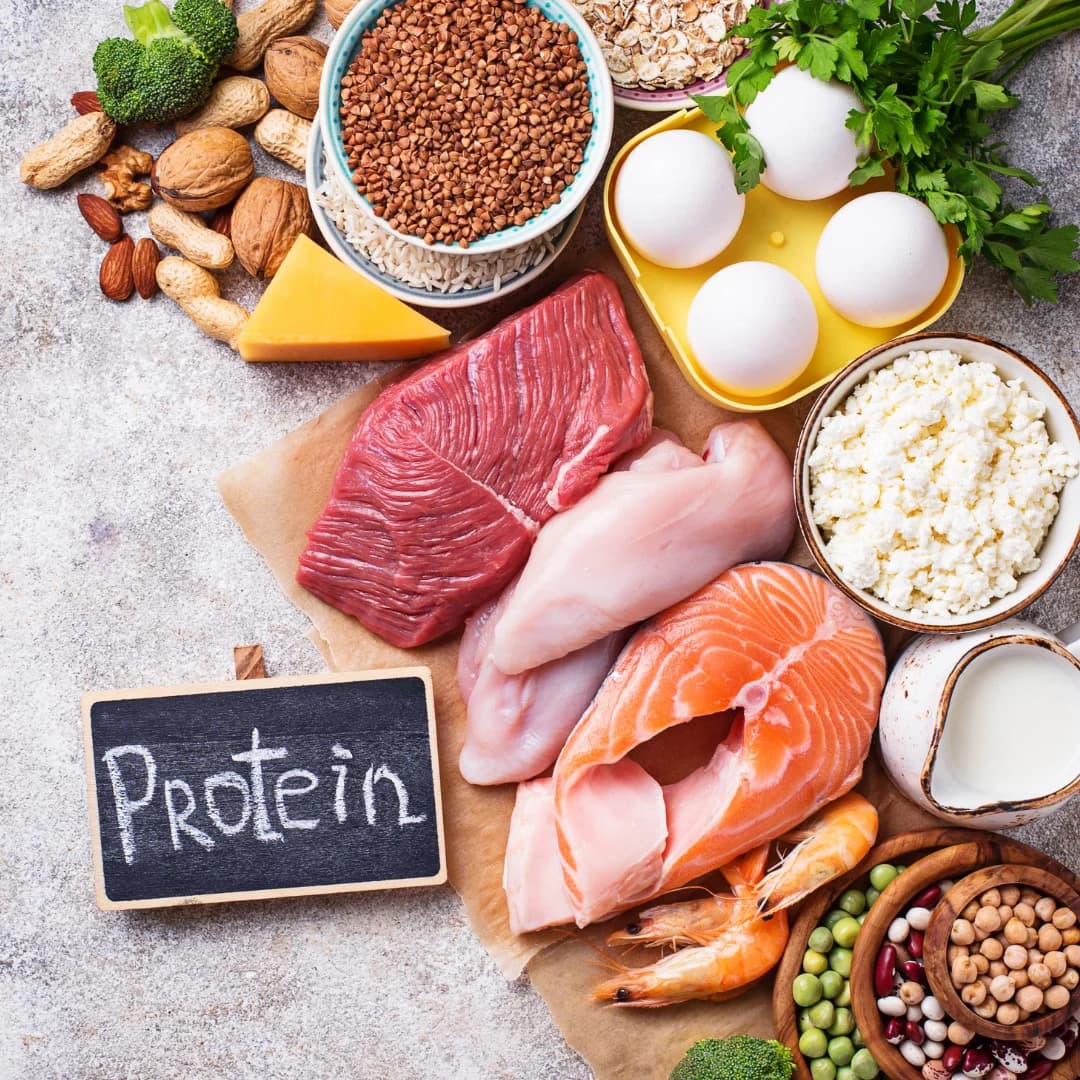 A variety of high-protein foods including raw salmon, beef, chicken, eggs, cottage cheese, shrimp, legumes, nuts, milk, and grains w