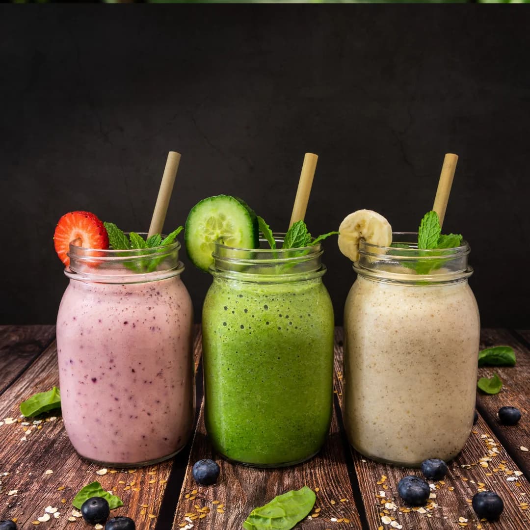 Three smoothies in mason jars with paper straws, garnished with cucumber, banana, strawberry, and mint on a rustic wooden surface