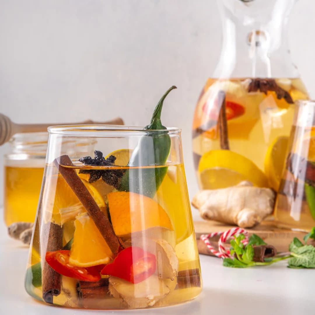 front view of healthy drink, green chili, ginger, cinnamon stick, cloves, orange slices, lemon in water served in glass also glass jug of same water in background