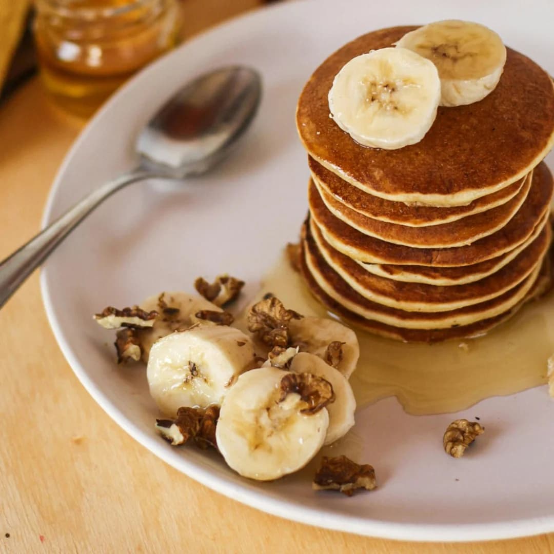 A tall stack of banana pancakes topped with banana slices, served with walnuts and syrup on a white plate with a spoon on the side.