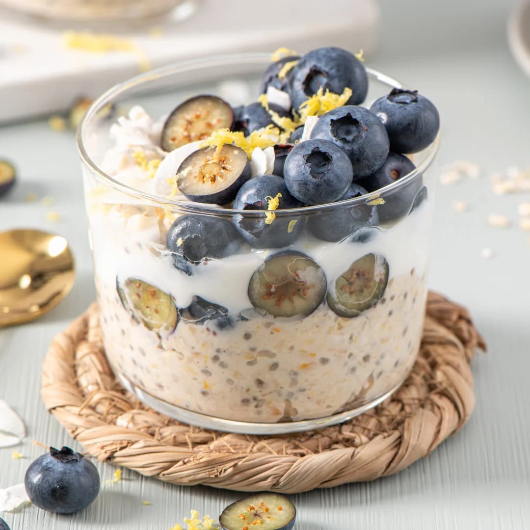 Glass jar of overnight oats layered with blueberries, yogurt, lemon zest, and chia seeds, placed on a woven coaster.