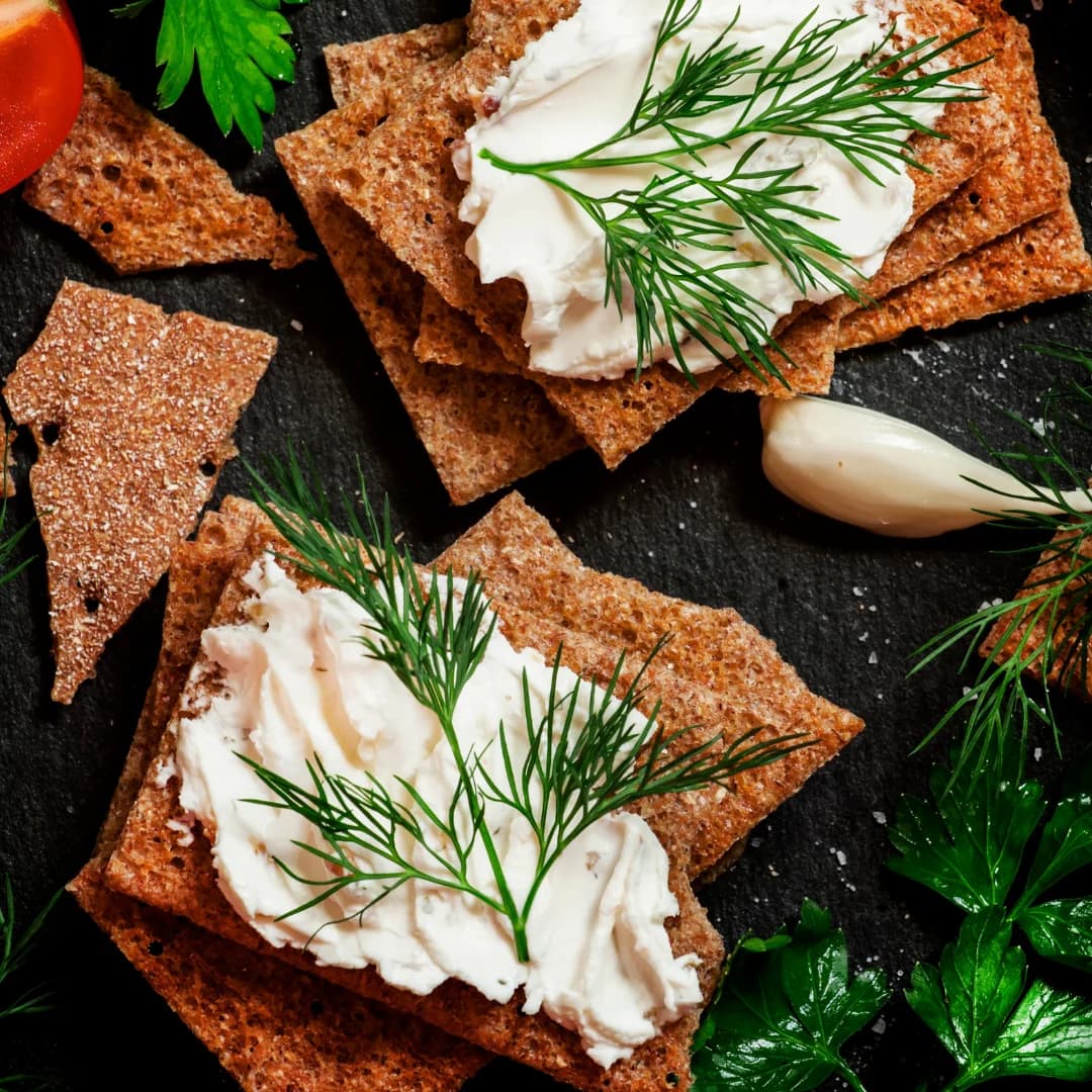 Crispbread topped with creamy cheese and fresh dill on a dark slate surface with herbs and garlic.