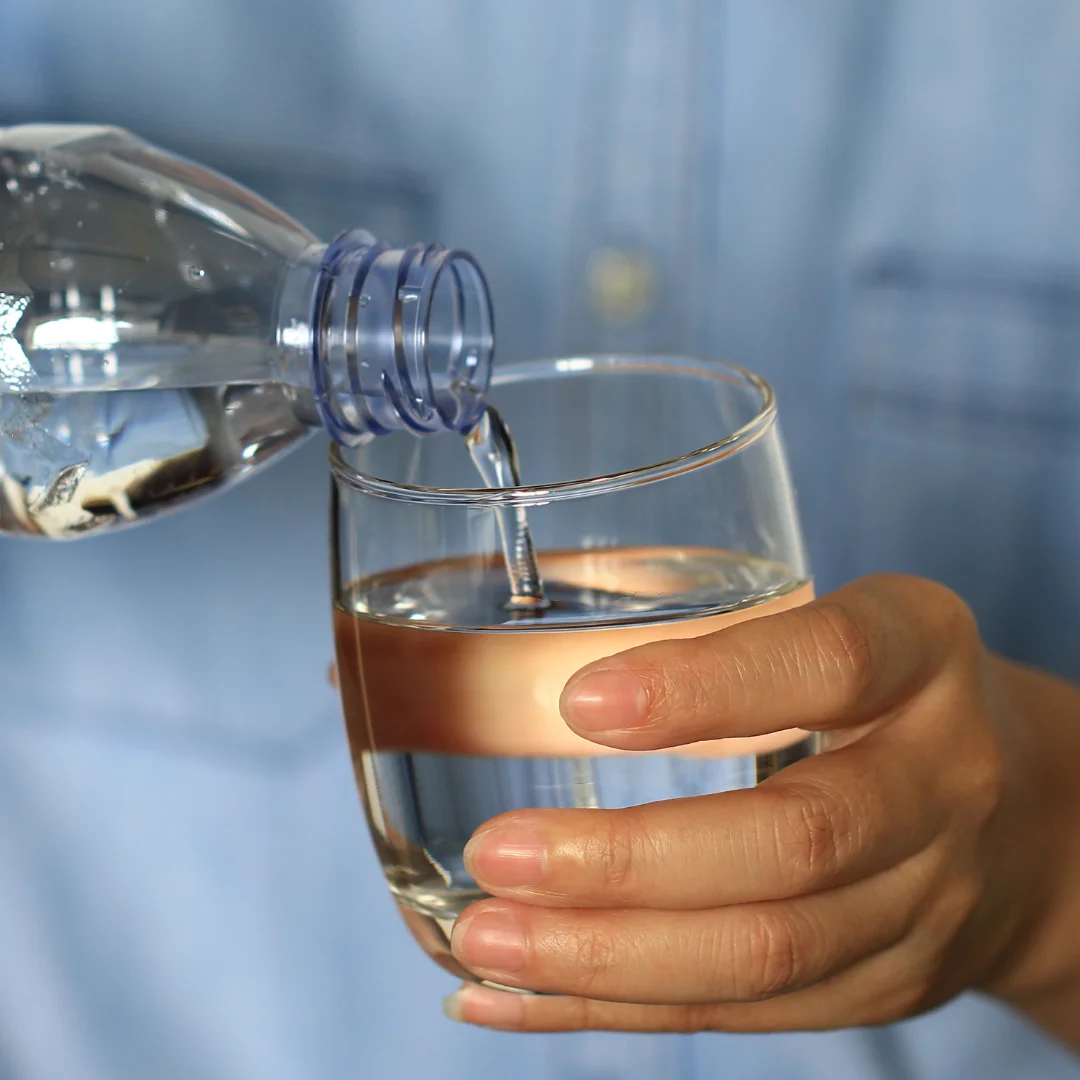 Hand pouring water from a plastic bottle into a clear glass, with a person in a blue shirt in the background