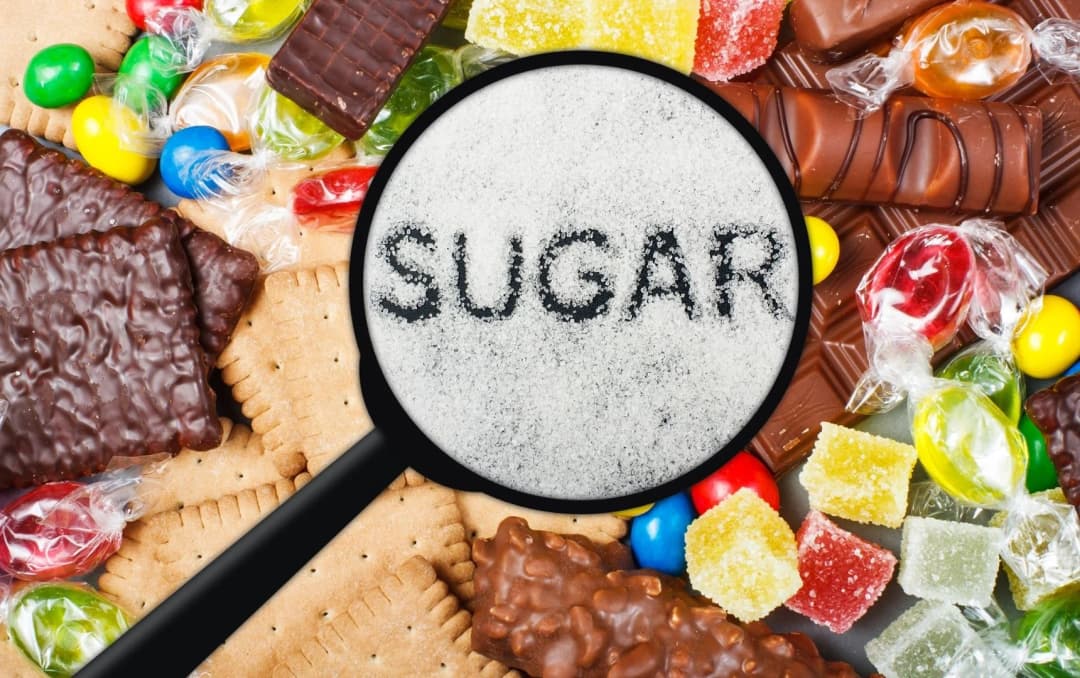 Magnifying glass showing the word sugar surrounded by candies, biscuits, and chocolates highlighting sugar dangers
