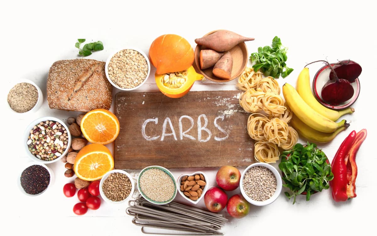 Assortment of healthy carbohydrate foods such as whole grains, pasta, sweet potatoes, fruits, and vegetables surrounding a wooden board