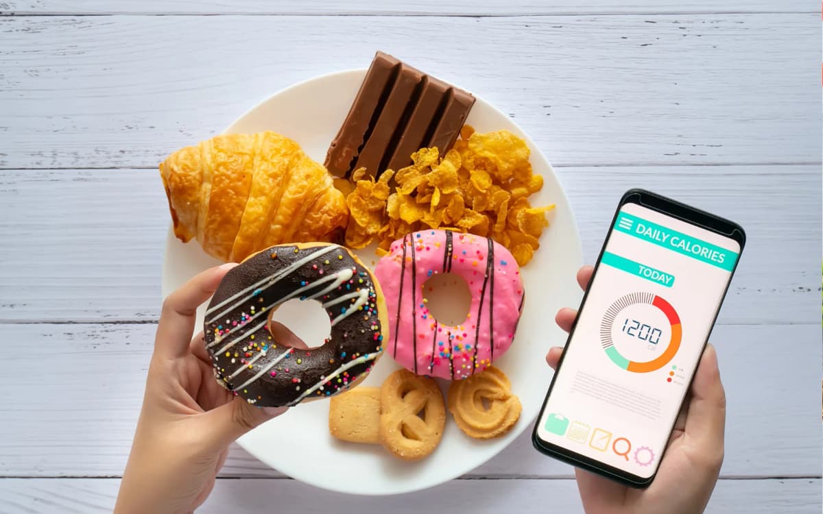 hand hollding donut for calculating calories on mobile and food plate on the table