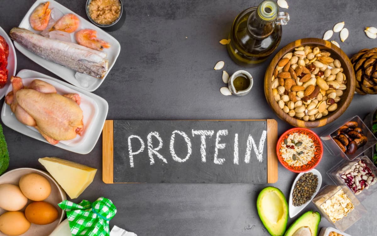 Assorted protein sources including raw chicken, fish, shrimp, eggs, nuts, seeds, avocado, cheese, grains, and a bottle of oil.