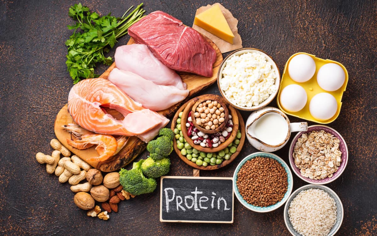 Protein sources displayed on a rustic background including salmon, beef, chicken, eggs, cheese, milk, legumes, grains and broccoli.
