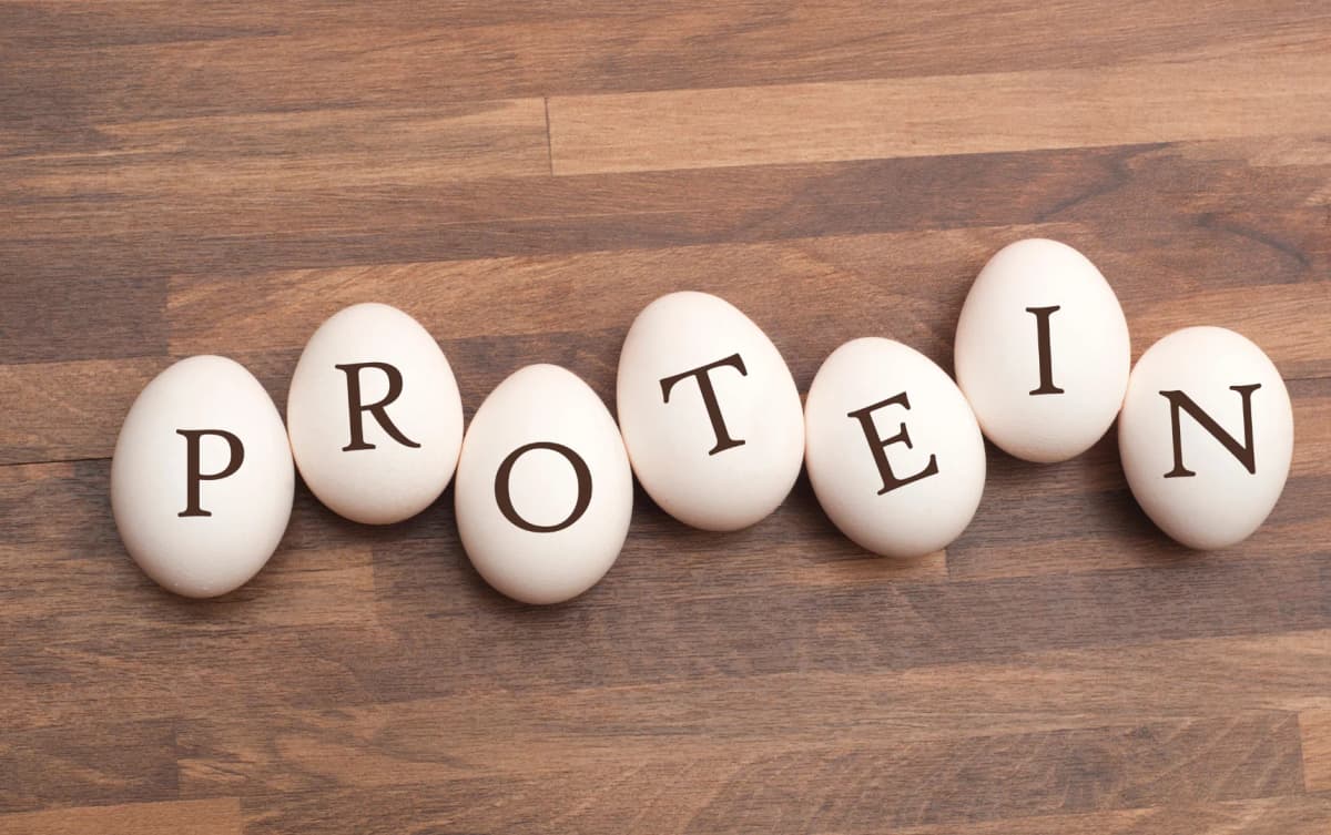 Seven white eggs lined up on a wooden surface, each with one letter of the word 'PROTEIN' printed on them.