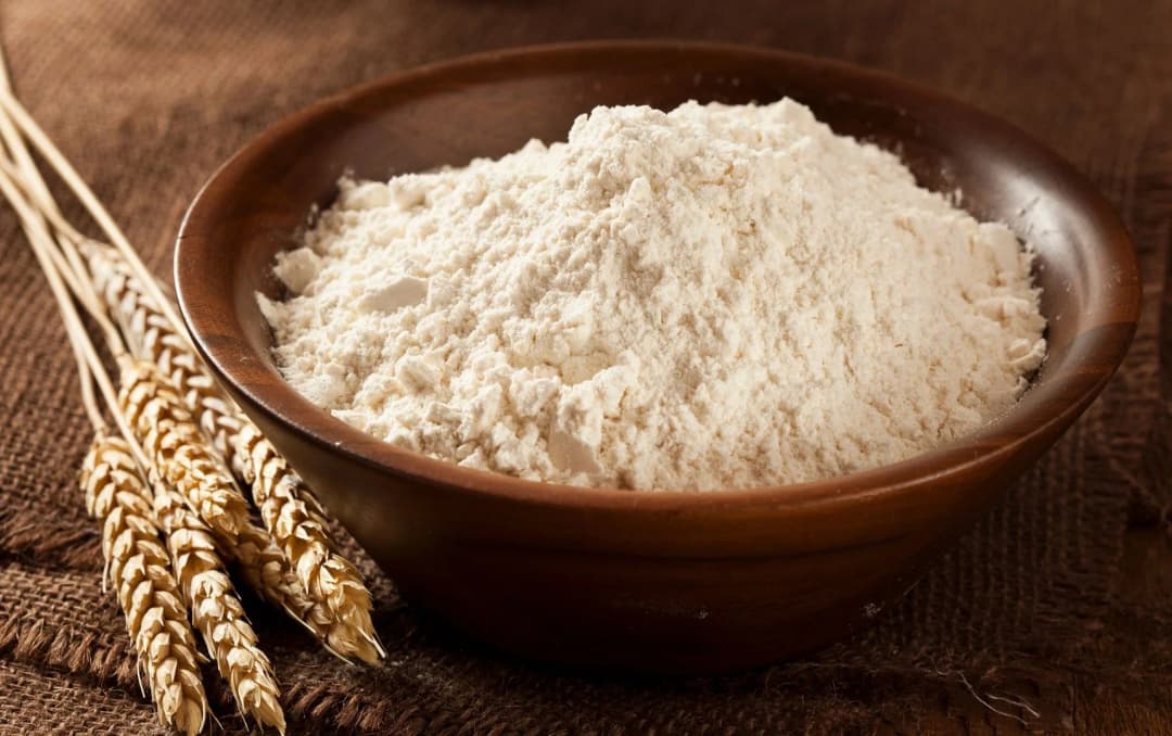 Bowl of white wheat flour with wheat stalks on a rustic brown cloth, healthy cooking ingredient