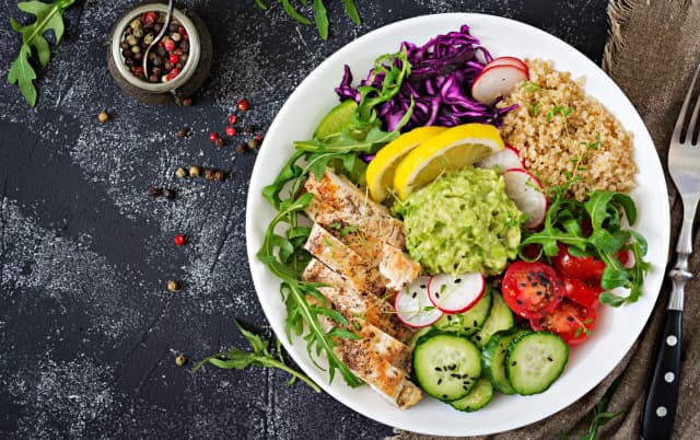Healthy chicken power bowl with grilled chicken, quinoa, guacamole, cherry tomatoes, cucumber, red cabbage, arugula, and lemon slices