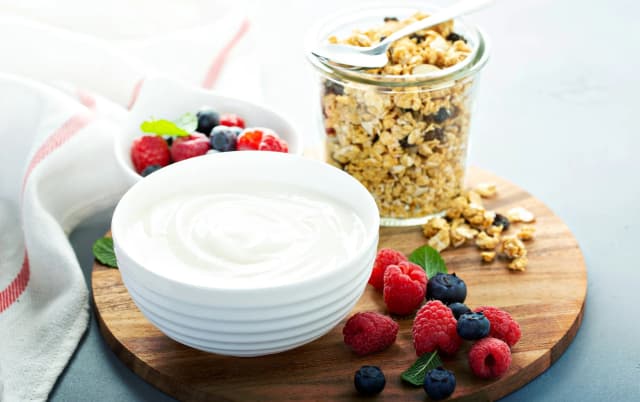 a bowl of greek yogurt served in white bowl place in rounded cutting board nut jar and cherry and strawberry in background