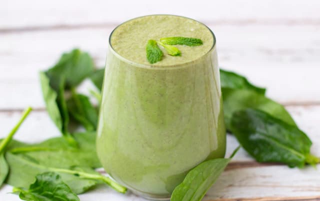 Green spinach smoothie in a clear glass garnished with mint leaves, surrounded by fresh spinach.