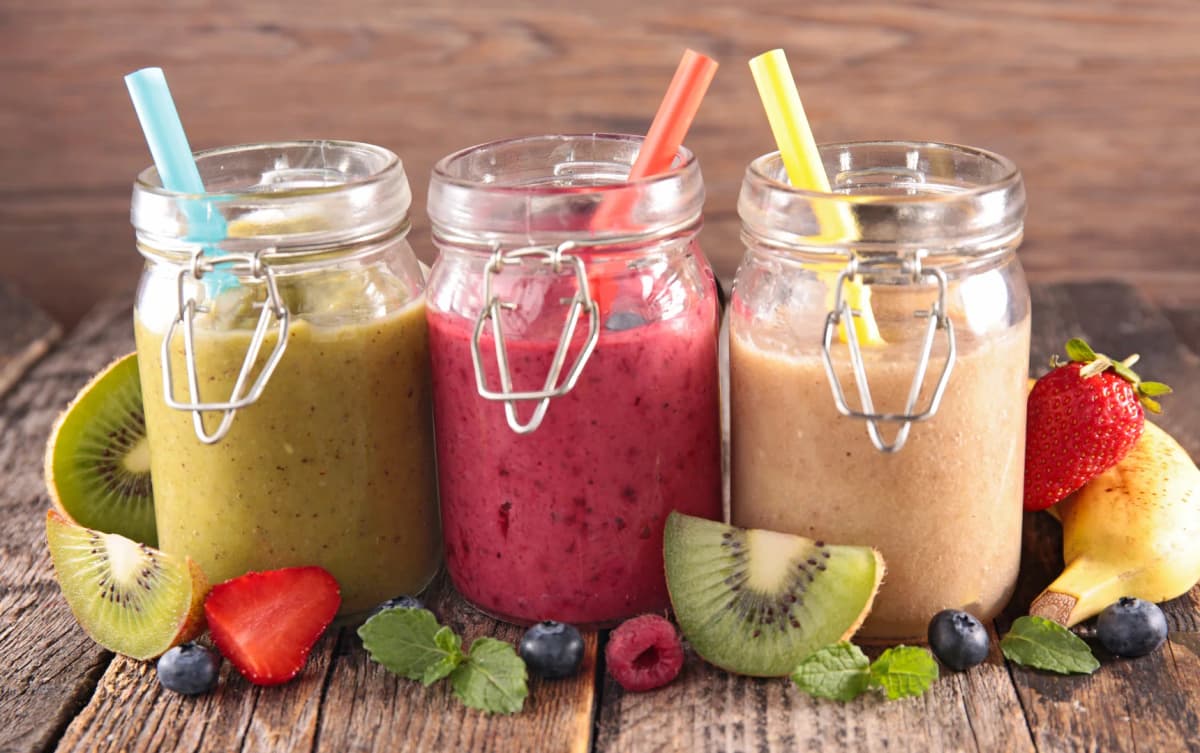 Three colorful smoothies in glass jars with straws, surrounded by fresh kiwi, strawberries, blueberries, and mint.