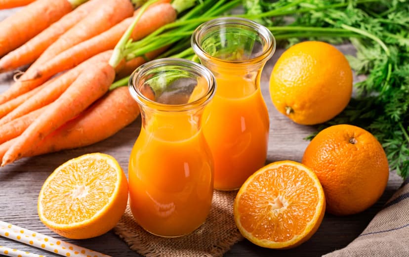 front view of carrot and orange juice in two glass jar have some carrot and oranges in background placed in rustic cloth