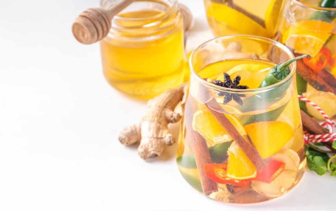top view of healthy drink, green chili, ginger, cinnamon stick, cloves, orange slices, lemon in water served in glass also glass jug of same water in background