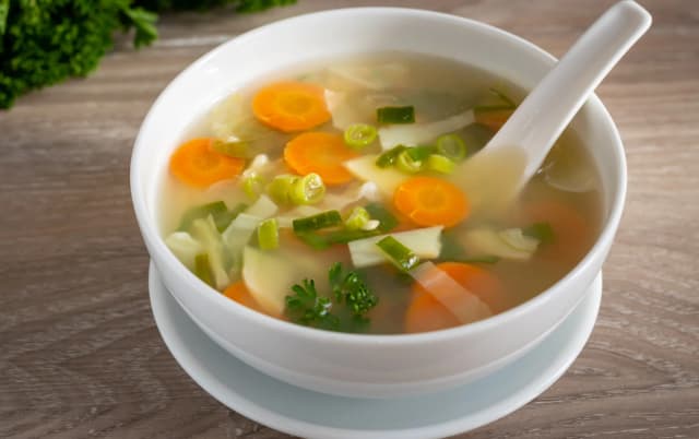 front view of carrot, green onion, ginger soup served in white bowl placed in wooden table