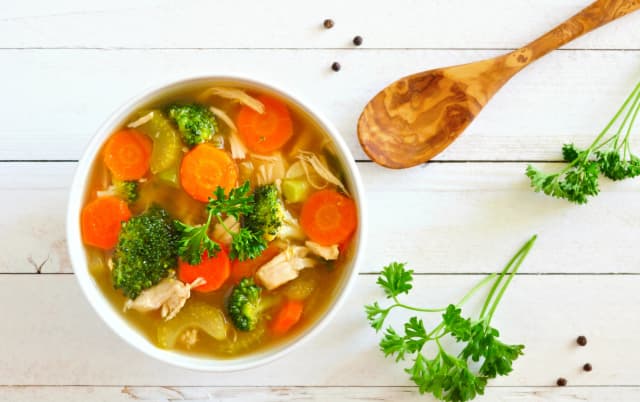 top view of spicy vegitable soup , brockly, carrot slices, cabbage, tomato, onion inside served in white bowl curriander and wooden spoon on the background