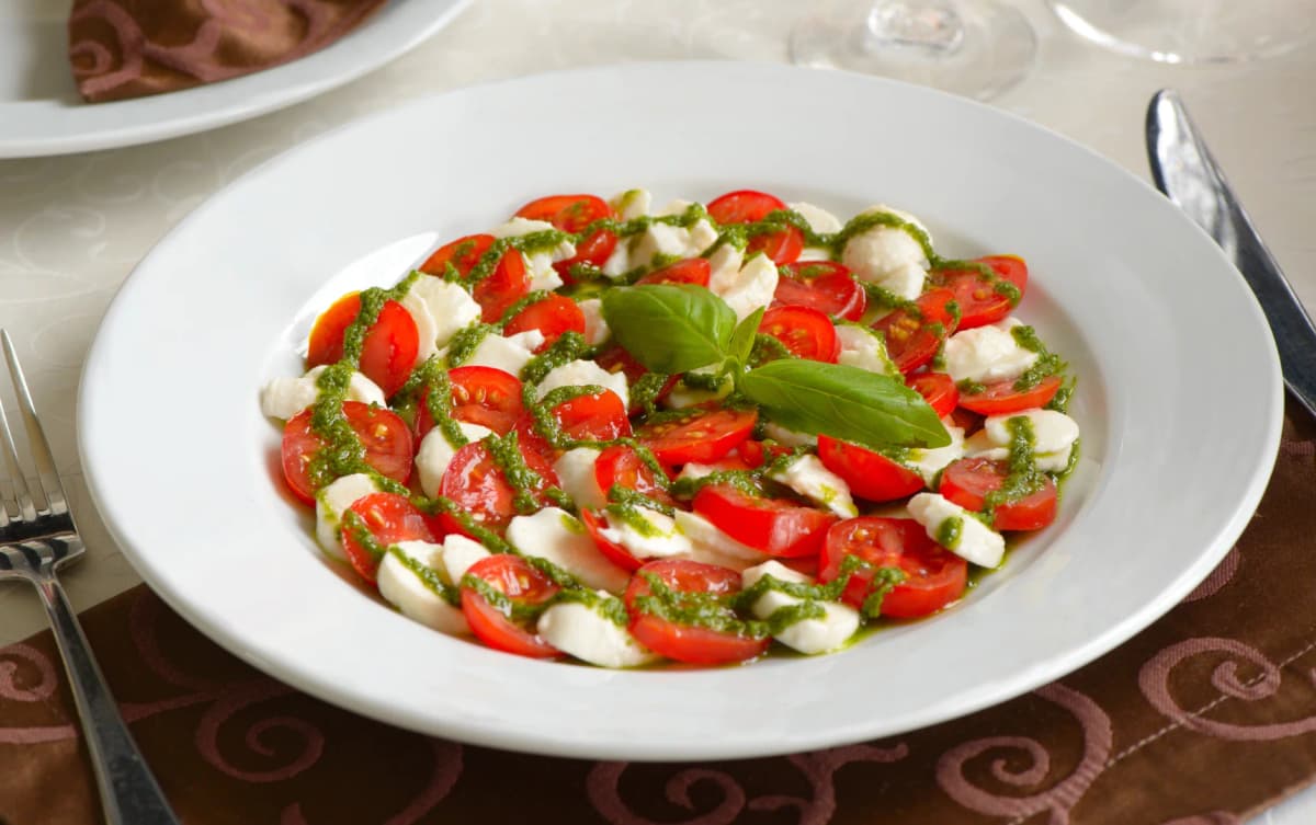 Caprese salad made with halved cherry tomatoes and mozzarella balls on a white plate, topped with green pesto and garnished with a basil leaf.