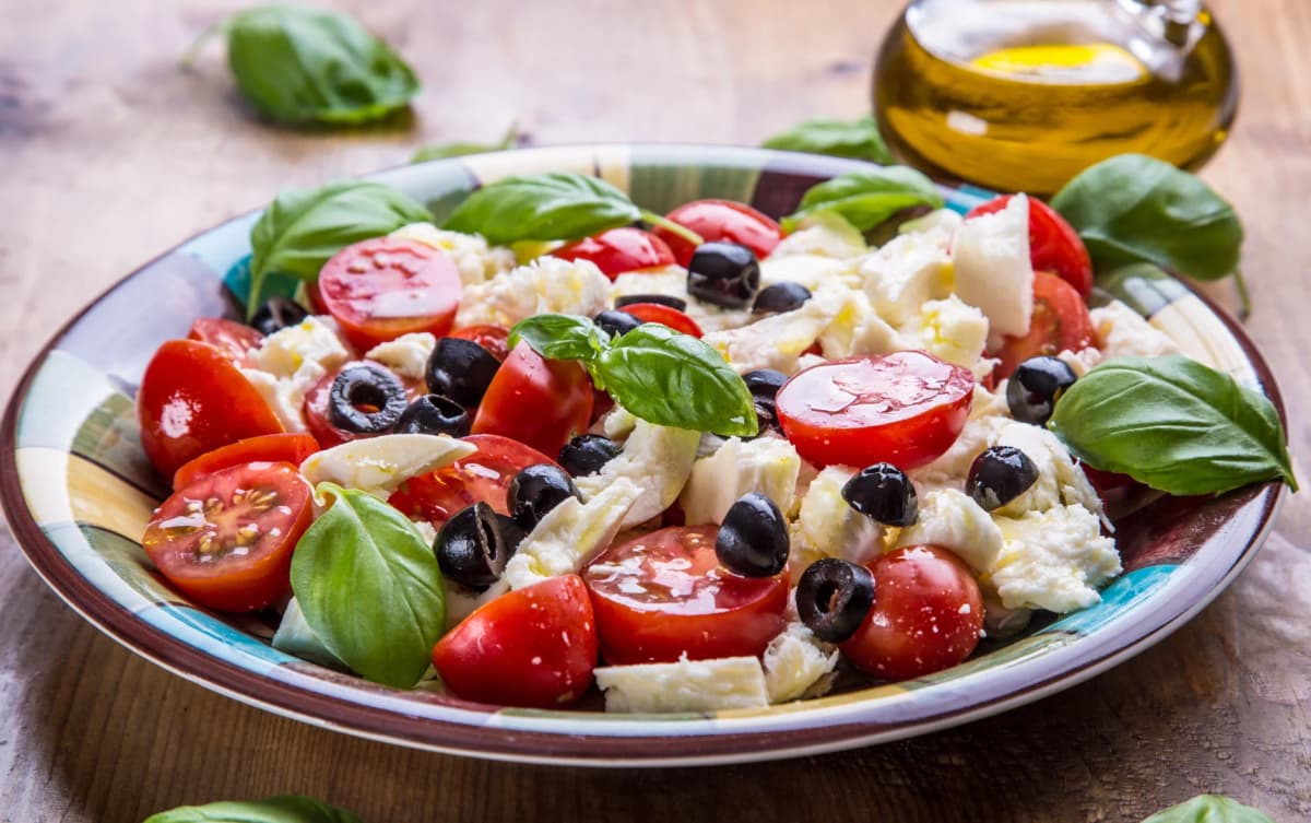 Caprese-style salad with cherry tomatoes, shredded mozzarella, black olives, and fresh basil leaves served in a colorful ceramic bowl.