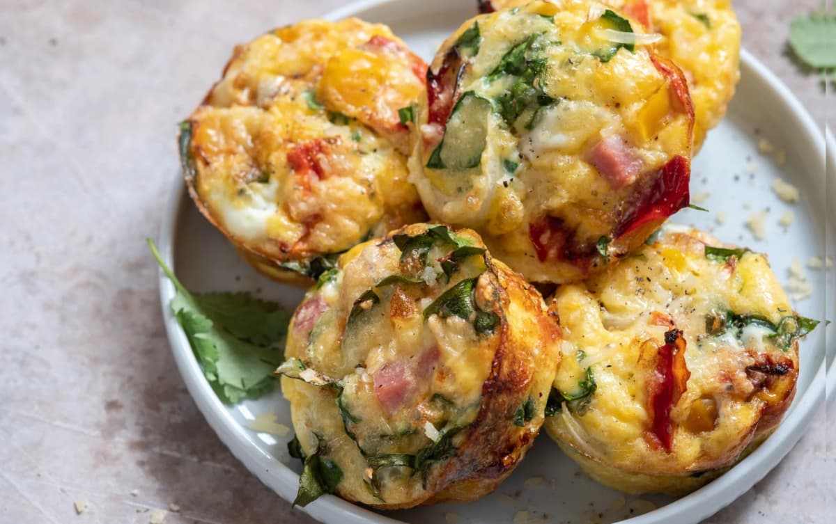 Cheesy egg muffins filled with bell peppers, ham, spinach, and herbs, served on a white dish with a garnish of cilantro.