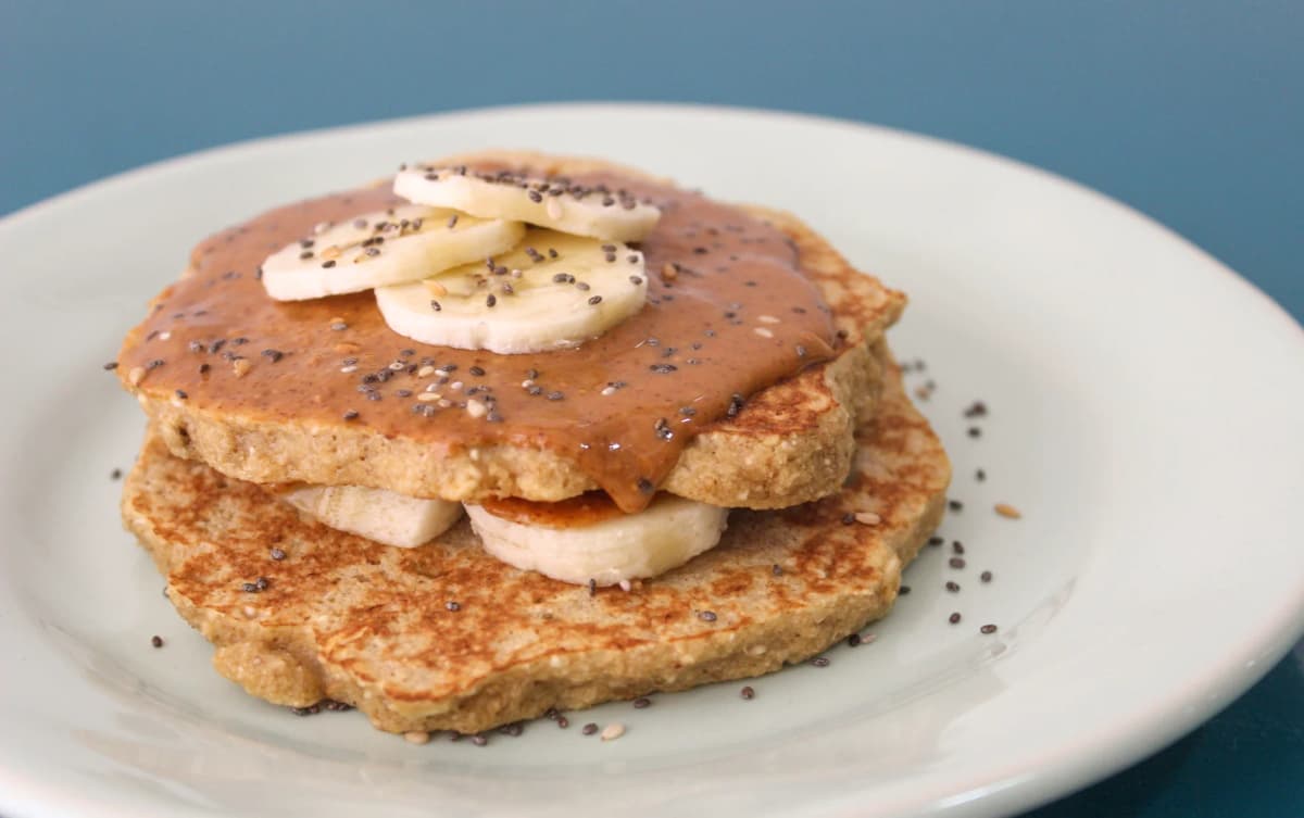 Banana pancakes topped with peanut butter, banana slices, and sprinkled chia and sesame seeds on a white plate.