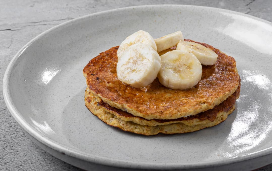 Two banana pancakes topped with banana slices, served on a plain grey plate with a light drizzle of syrup.