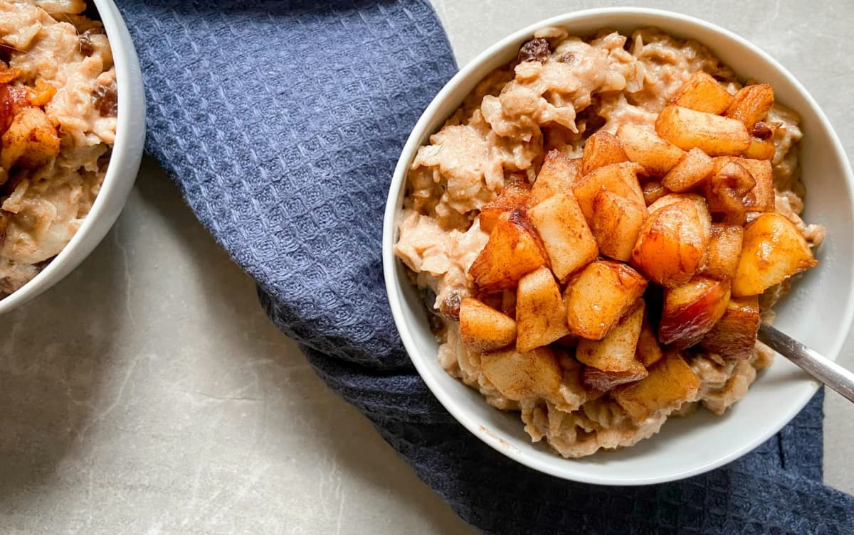 Bowl of cinnamon oatmeal topped with caramelized apple chunks, placed on a gray surface with a blue textured napkin.