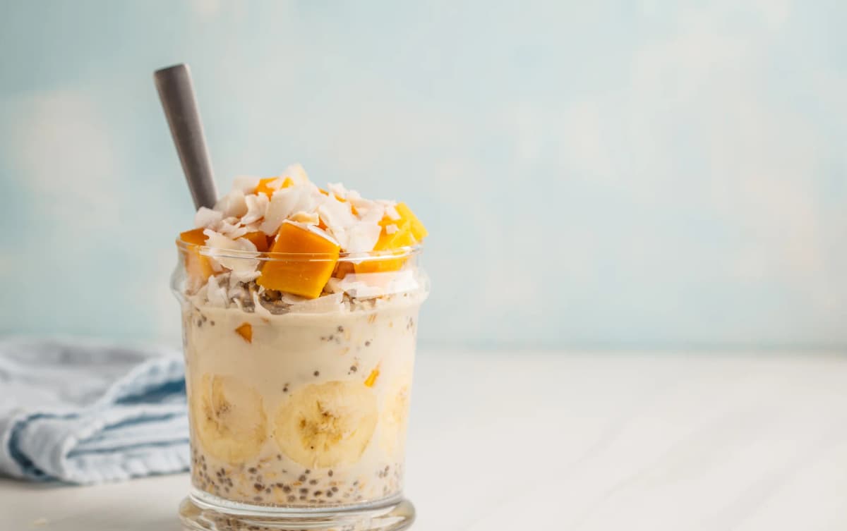Glass jar of overnight oats with banana slices, mango chunks, coconut flakes, and a spoon inside.