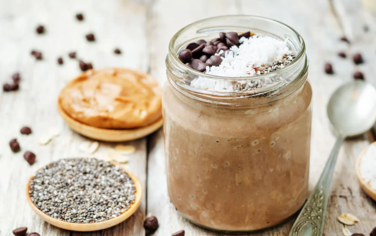 Jar of chocolate overnight oats topped with shredded coconut, chia seeds, and chocolate chips, with peanut butter in the background.