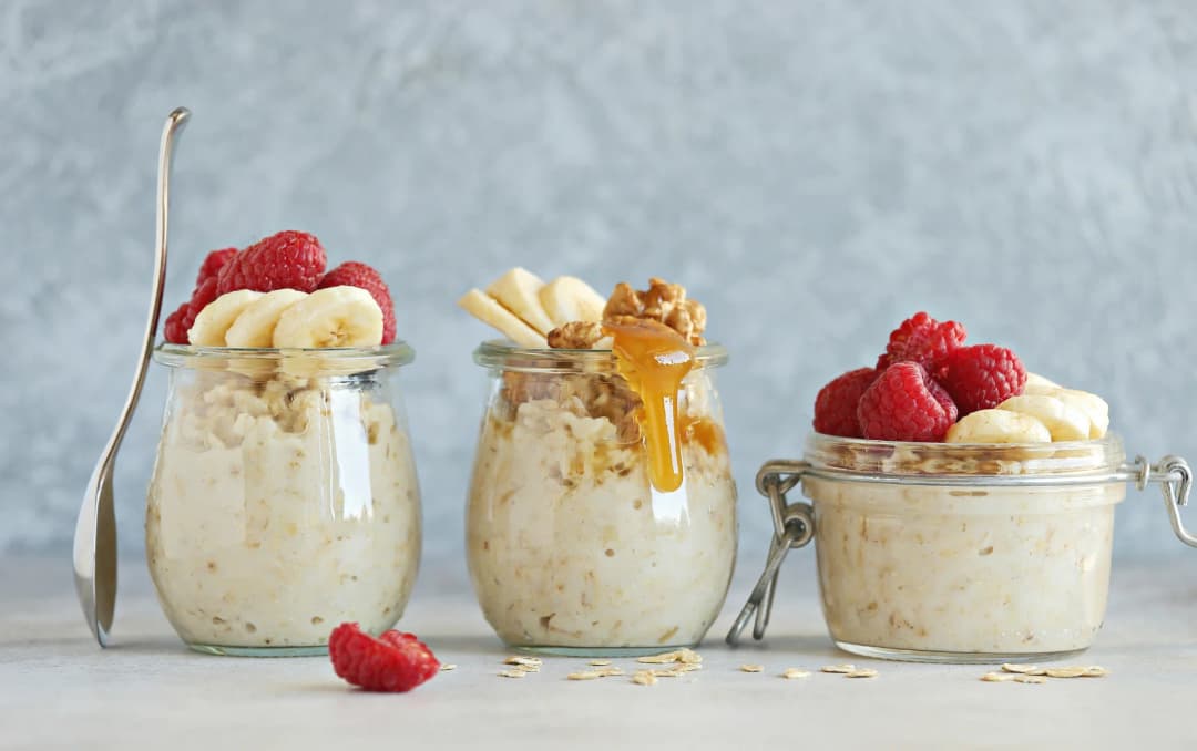 Three glass jars of overnight oats topped with banana slices, raspberries, honey, and granola against a light gray background.
