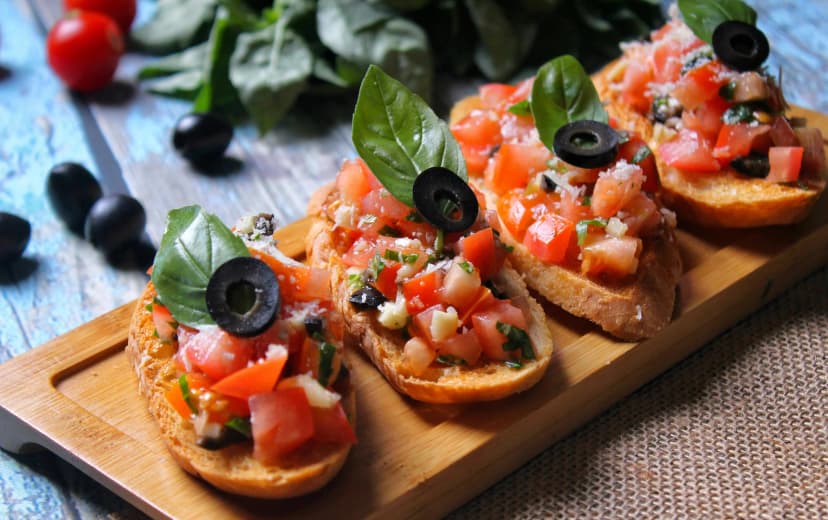 Top view of four bread toasts topped with tomato cubes, cheese pieces, and olive slices served on a wooden board.