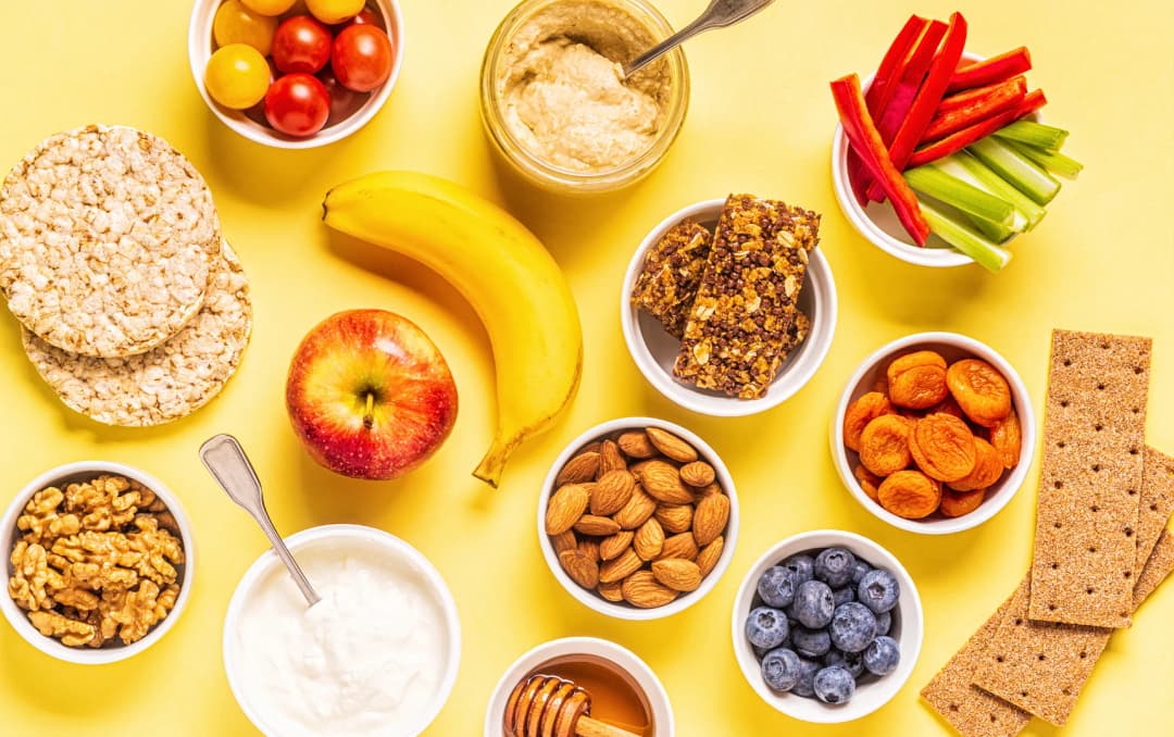 top view lots of healthy snacks like almond, apple, banana, carrot, cucumber, rice cake, yogurt and more