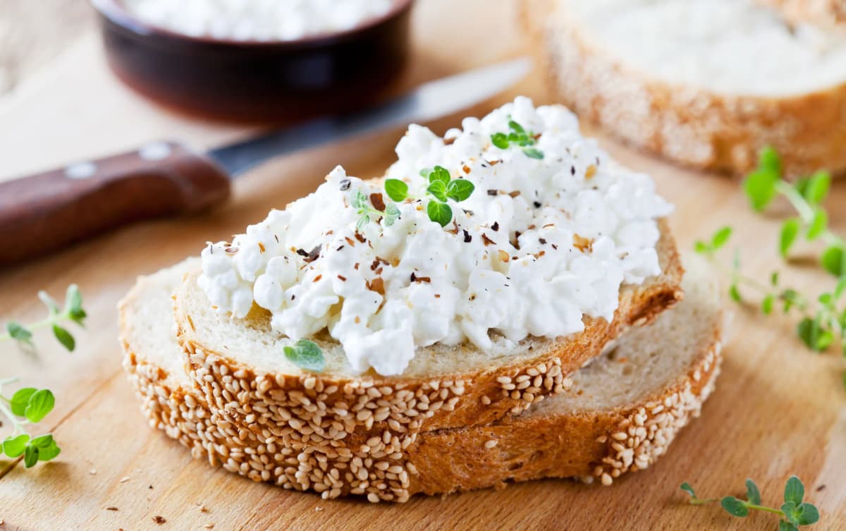 Sesame seed bread slices topped with cottage cheese, black pepper, and fresh oregano on a wooden board.