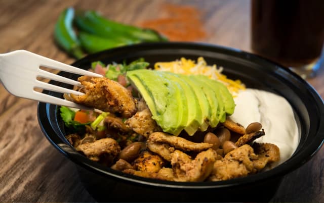a grilled chicken with slices of avocado served in the black bowl green chili on the background placed in wooden table