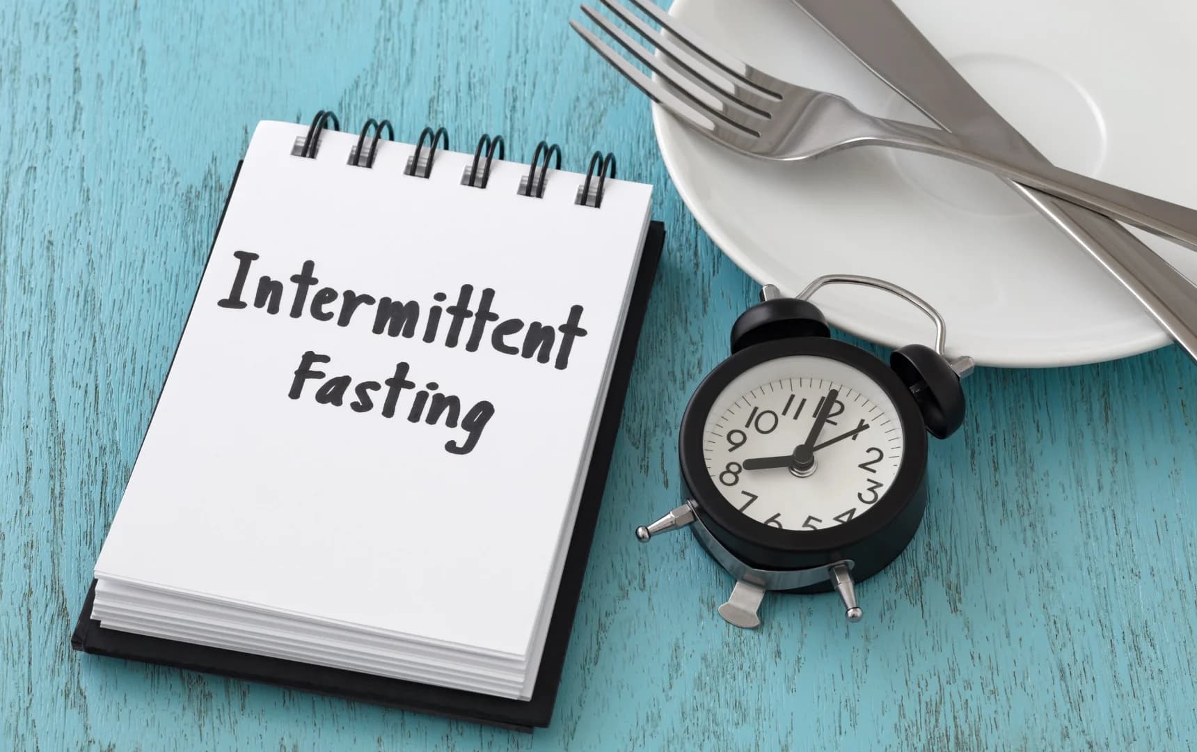 Notebook with text Intermittent Fasting beside plate, cutlery and alarm clock