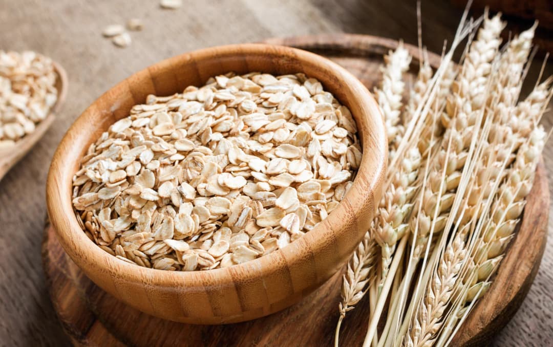 Wooden bowl filled with rolled oats next to wheat stalks on a rustic table