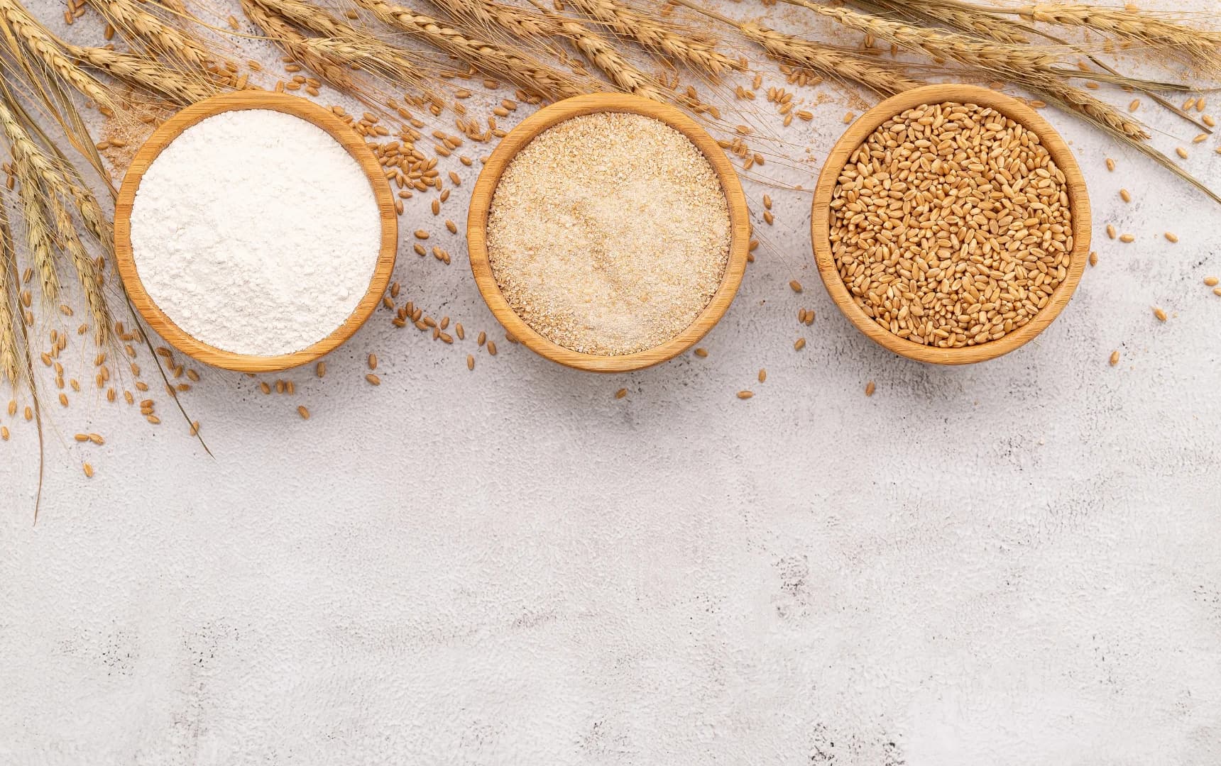Wheat products in wooden bowls – whole wheat grains, semolina, and flour on a light background with wheat stalks
