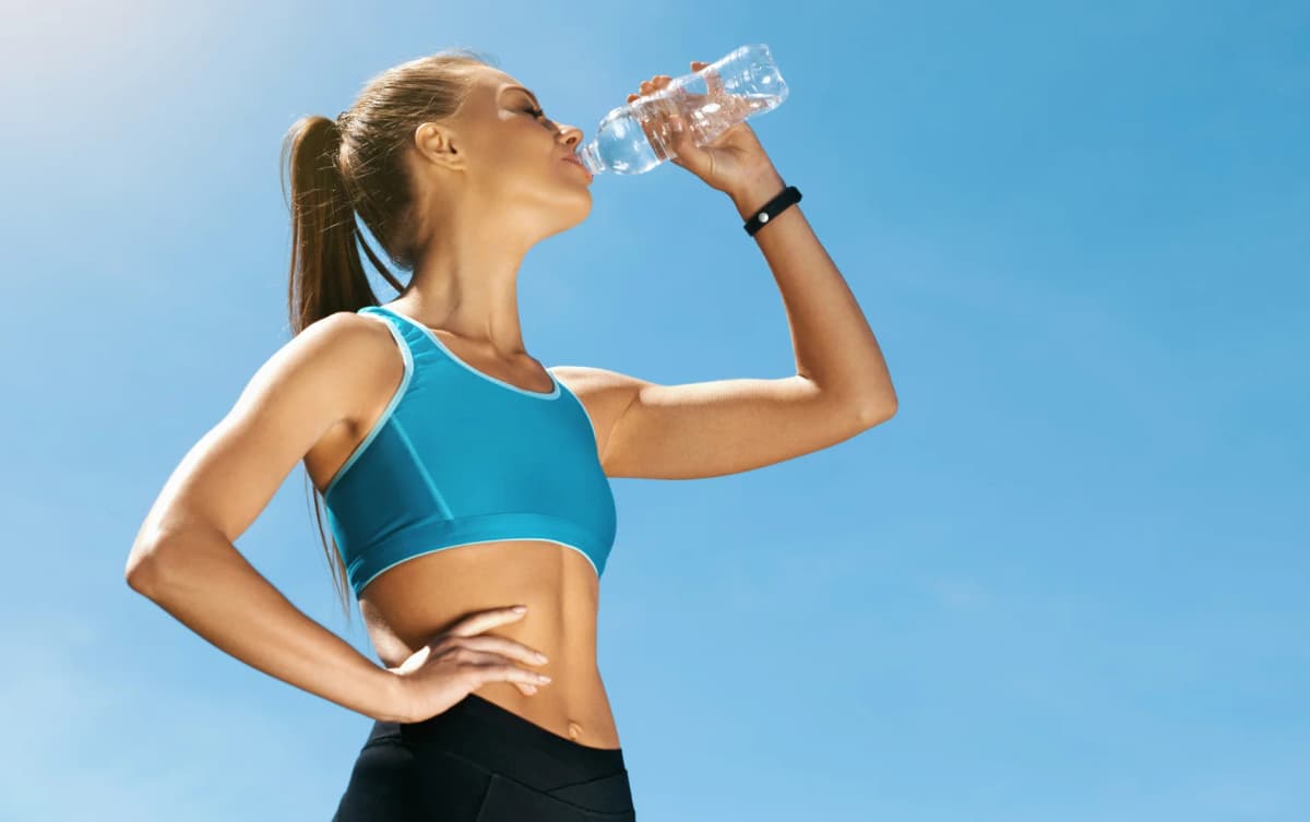 Fit young woman in athletic wear drinking water from a plastic bottle under a bright blue sky.