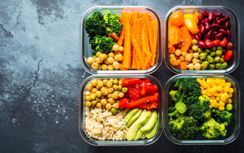Colorful meal prep containers filled with fresh vegetables, chickpeas, quinoa, and legumes arranged on a dark background