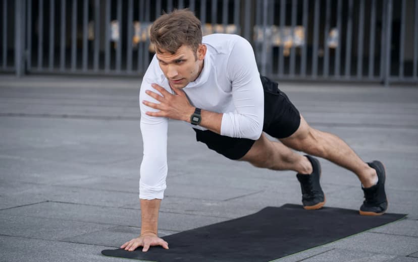 Man doing a shoulder tap plank exercise outdoors on a black mat, wearing a white long-sleeve shirt and black shorts.