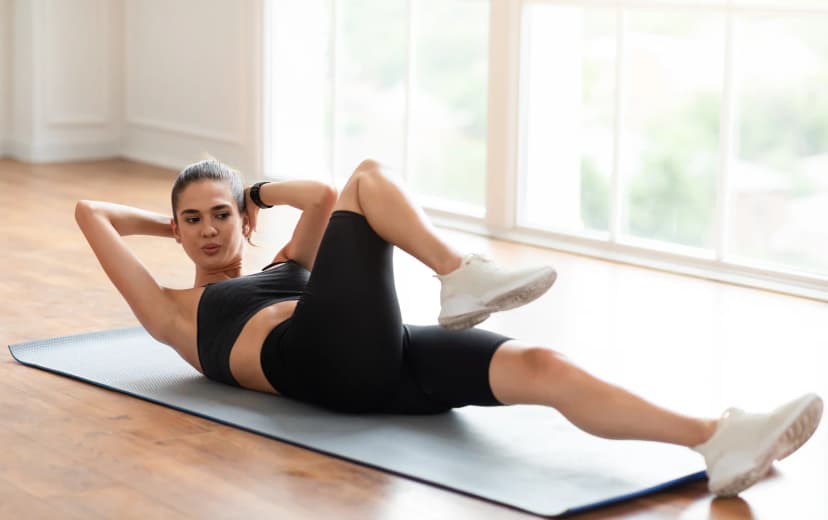 Woman performing a bicycle crunch exercise on a yoga mat in a bright indoor space, wearing black workout clothes.