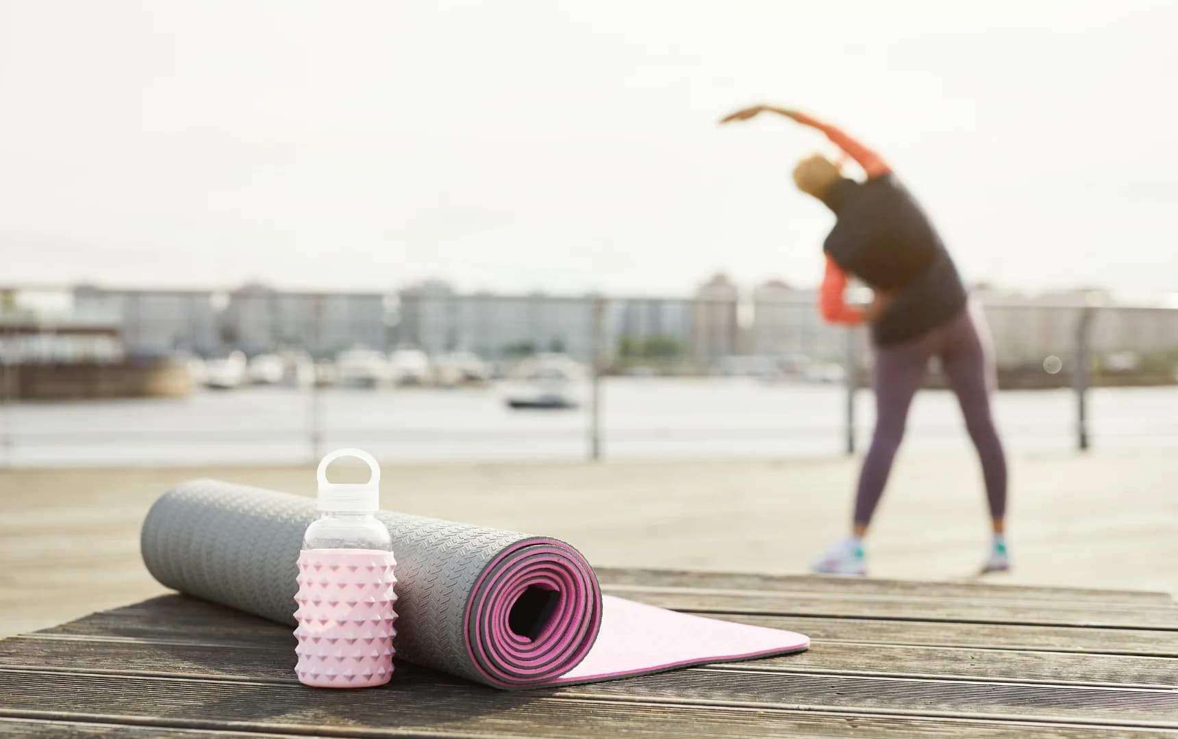 Close-up of a rolled yoga mat and pink water bottle on a wooden surface, with a woman stretching near a waterfront in the blurred background.