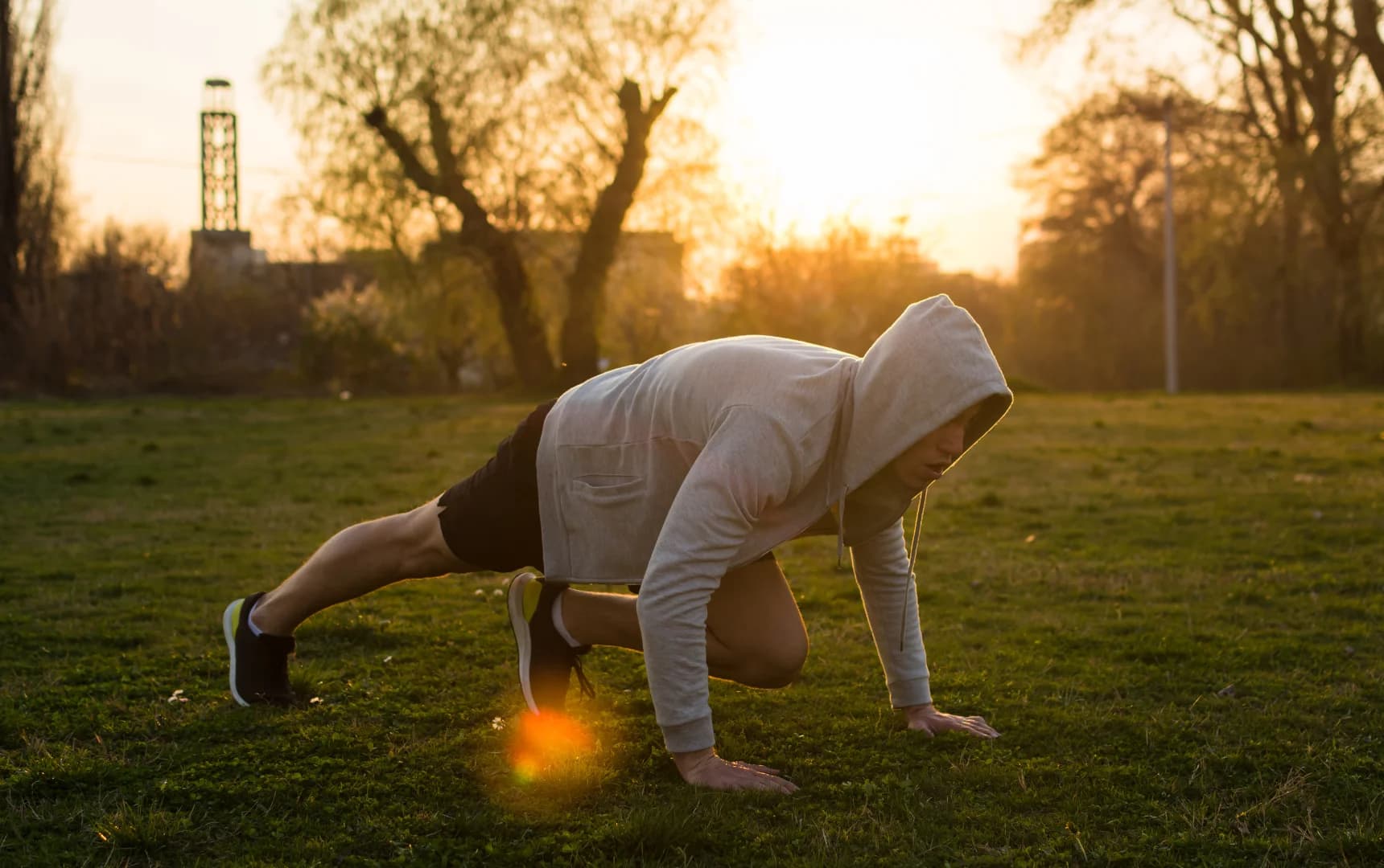 Man wearing a hoodie and shorts performing mountain climbers on grass at sunset in an outdoor park.