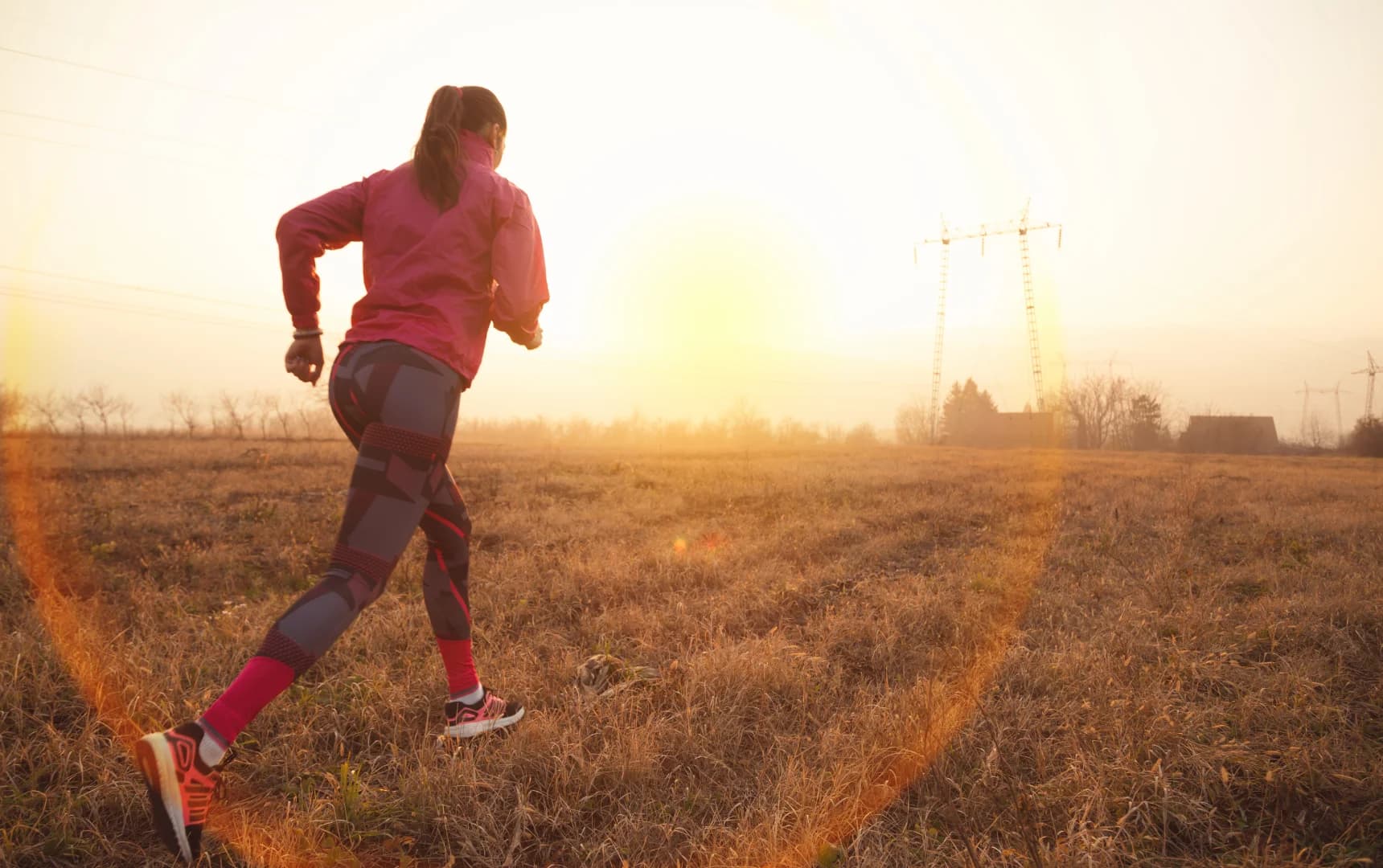 Woman in bright workout clothes running through a dry grassy field during sunrise or sunset.